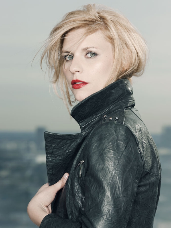 Close-up portrait of a woman with blonde hair wearing a black leather jacket against a blurred urban background, facing the camera.