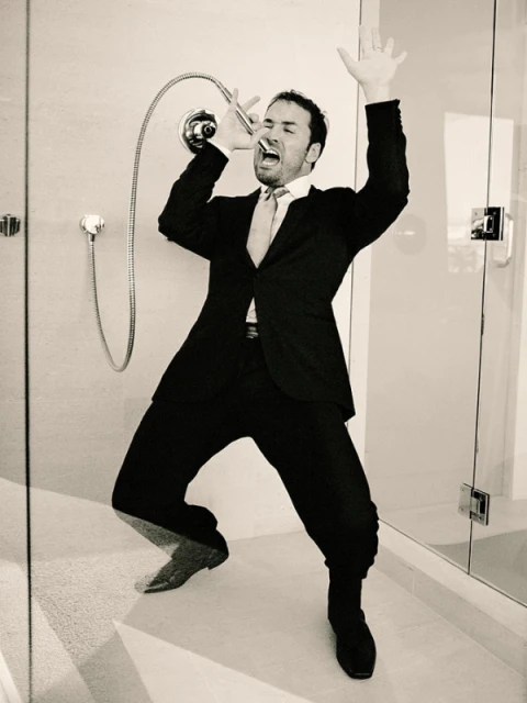 Man in a suit sings into a shower head in a tiled bathroom, arms raised, in a black and white photo.
