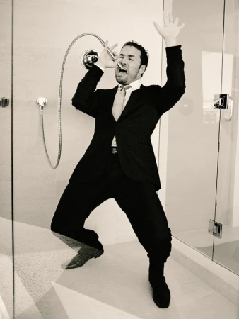 Man in a suit sings into a shower head in a tiled bathroom, arms raised, in a black and white photo.