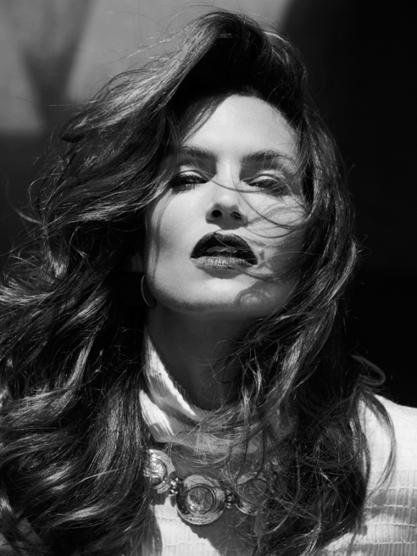 Black and white portrait of an adult woman with long hair, wearing a necklace, turtleneck sweater, and dark lipstick against a dark background.