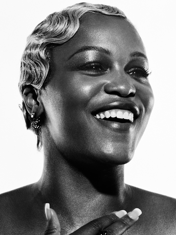 Monochrome portrait of a smiling, dark-skinned woman with short, waved hair, wearing earrings, and an elegant ring in a bright indoor setting.