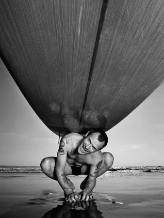 A tattooed man crouching on a wet sandy beach while carrying a large surfboard overhead, by Andrew Macpherson.