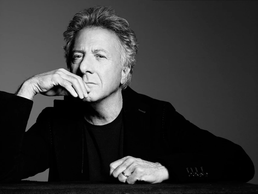 Black and white portrait of Dustin Hoffman in a dark jacket, resting his chin on his hand, posing against a grey background.