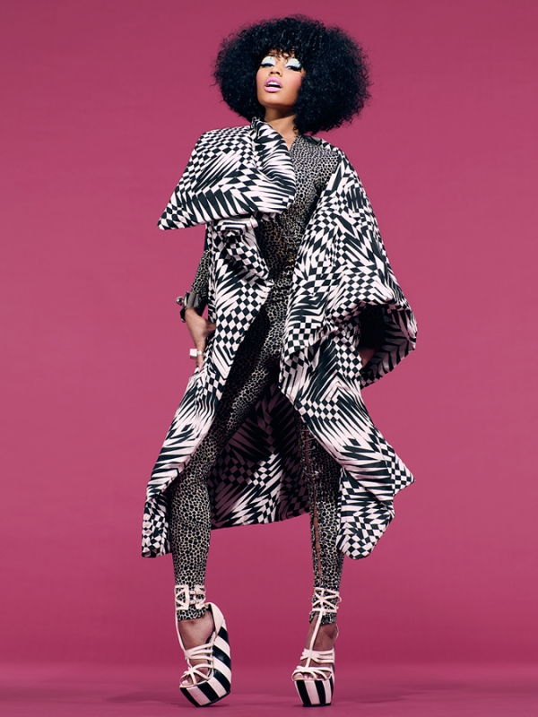 A woman with an afro stands in a patterned coat and leggings against a pink background in a fashion-forward studio shot.