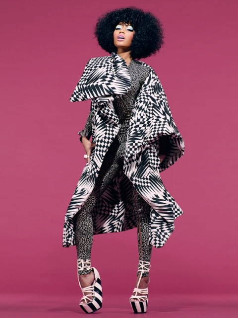 A woman with an afro stands in a patterned coat and leggings against a pink background in a fashion-forward studio shot.