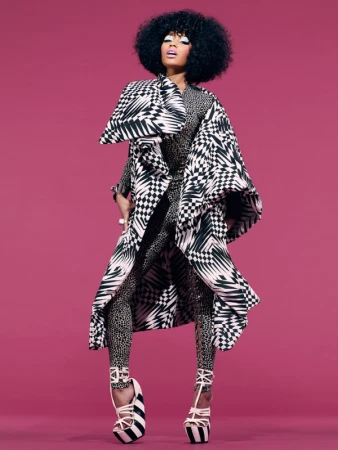 A woman with an afro stands in a patterned coat and leggings against a pink background in a fashion-forward studio shot.