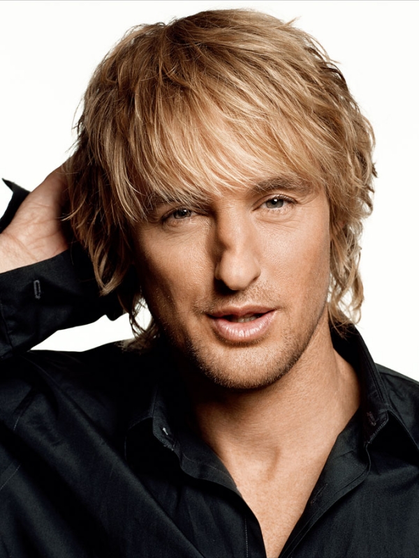 Portrait of Owen Wilson with messy blonde hair wearing a black button-down shirt against a solid white background