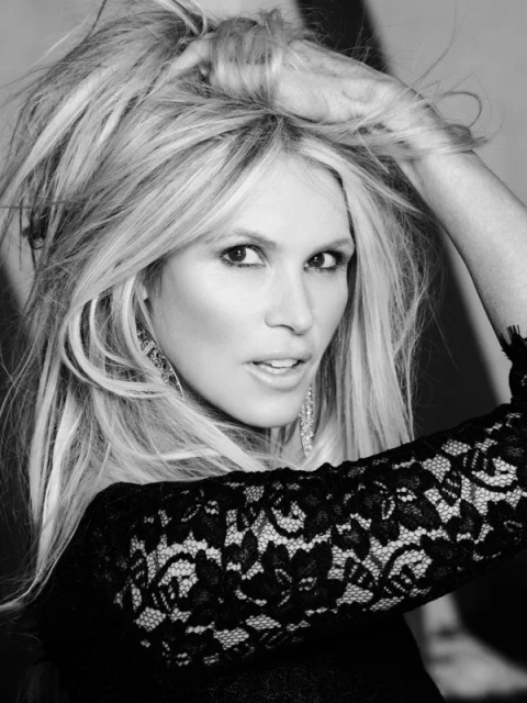 Black and white portrait of a blonde woman with her hand in her hair wearing lace top and earrings looking towards the camera indoors.