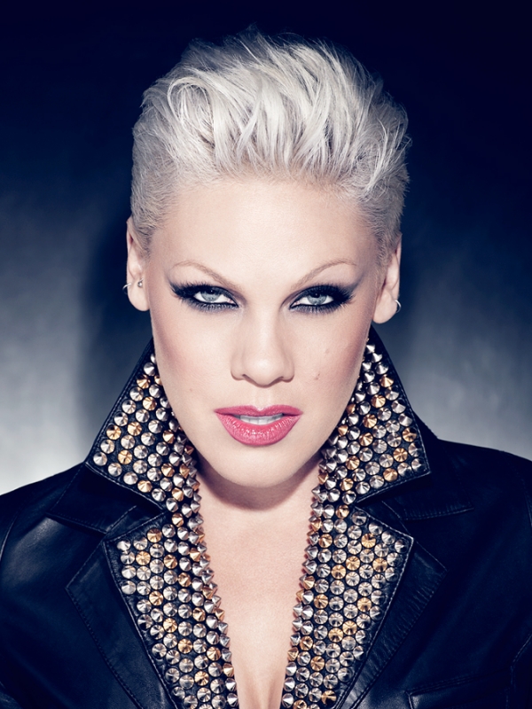 Close-up portrait of a woman with short, white hair and black leather jacket adorned with gold and silver studs, against a dark background.