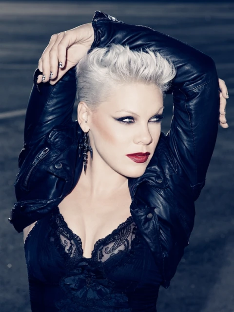 Close-up portrait of a woman with short platinum hair, dark makeup, and a black leather jacket, posing against a dark urban background.