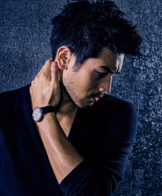 A man in a V-neck shirt and wristwatch poses against a textured dark gray background in a portrait-style fashion shot.