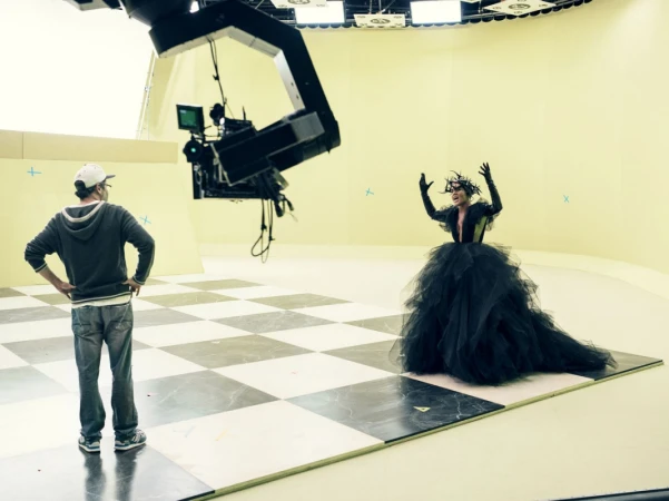 Behind-the-scenes shot featuring a performer in a dark, theatrical dress and a crew member in a film studio, complete with camera crane.