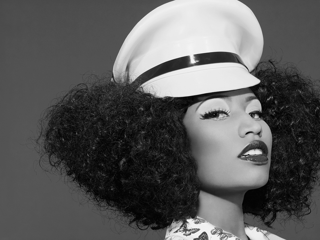 Black and white portrait of a woman with voluminous curly hair wearing a white peaked cap, posing in front of a dark gray backdrop.