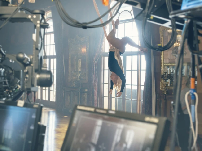 A blonde-haired woman in a black bodysuit performs aerial acrobatics indoors, suspended from fabric, framed by studio equipment and large windows.
