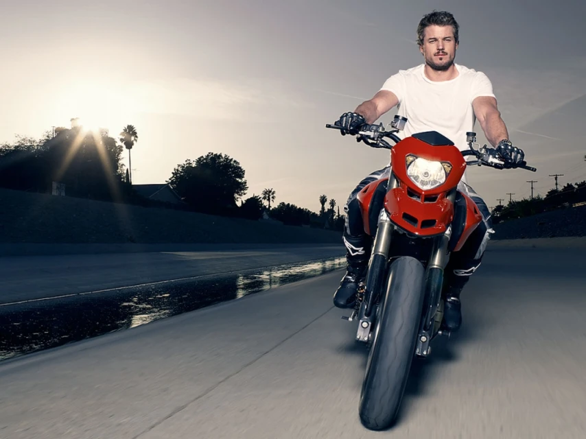 A man riding a red motorcycle on an asphalt road with sun flares in the background, wearing a white t-shirt and black riding gear.