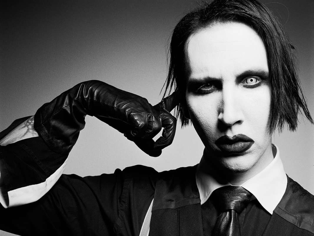 Monochrome portrait of Marilyn Manson in suit and leather gloves, holding a finger to his head, against a grey studio backdrop.