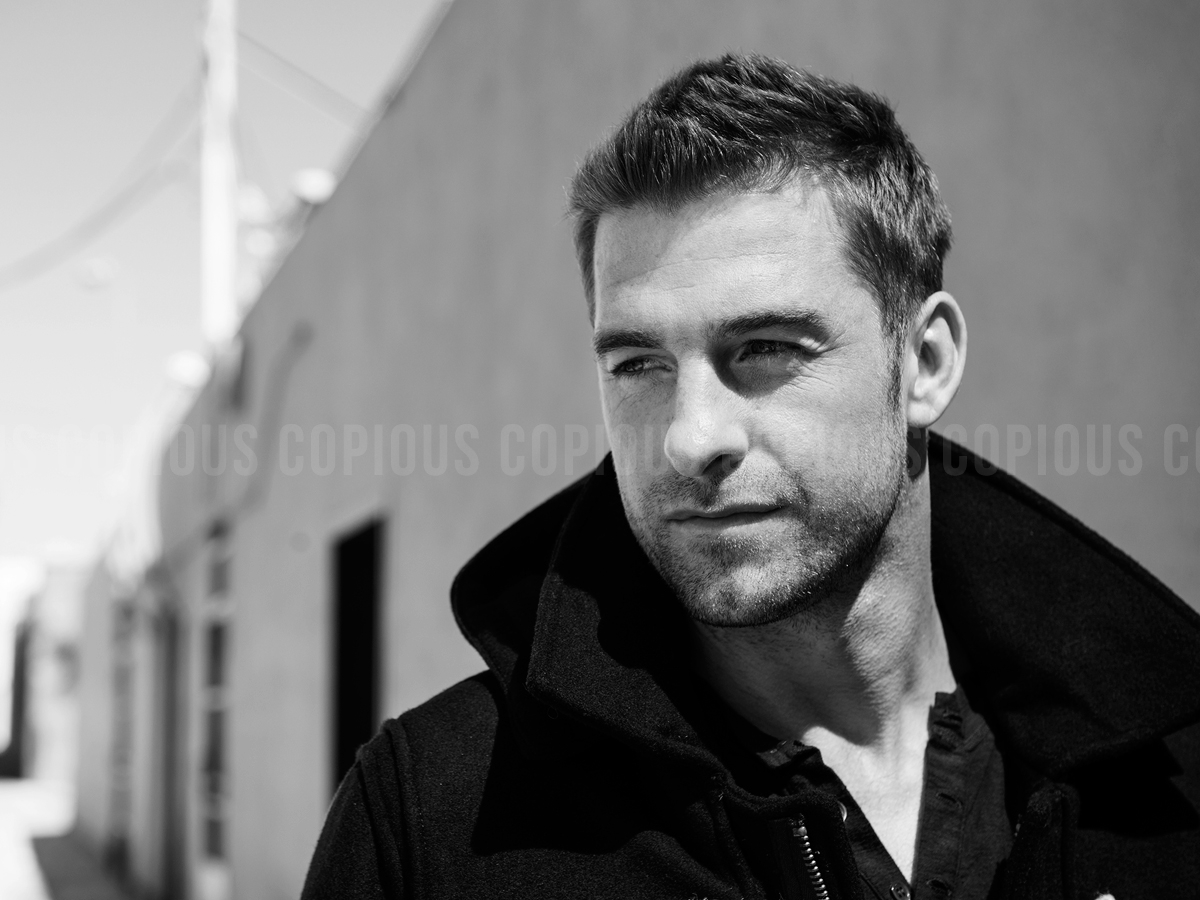 Black and white portrait of a man in a dark jacket with a high collar outdoors against a light building wall