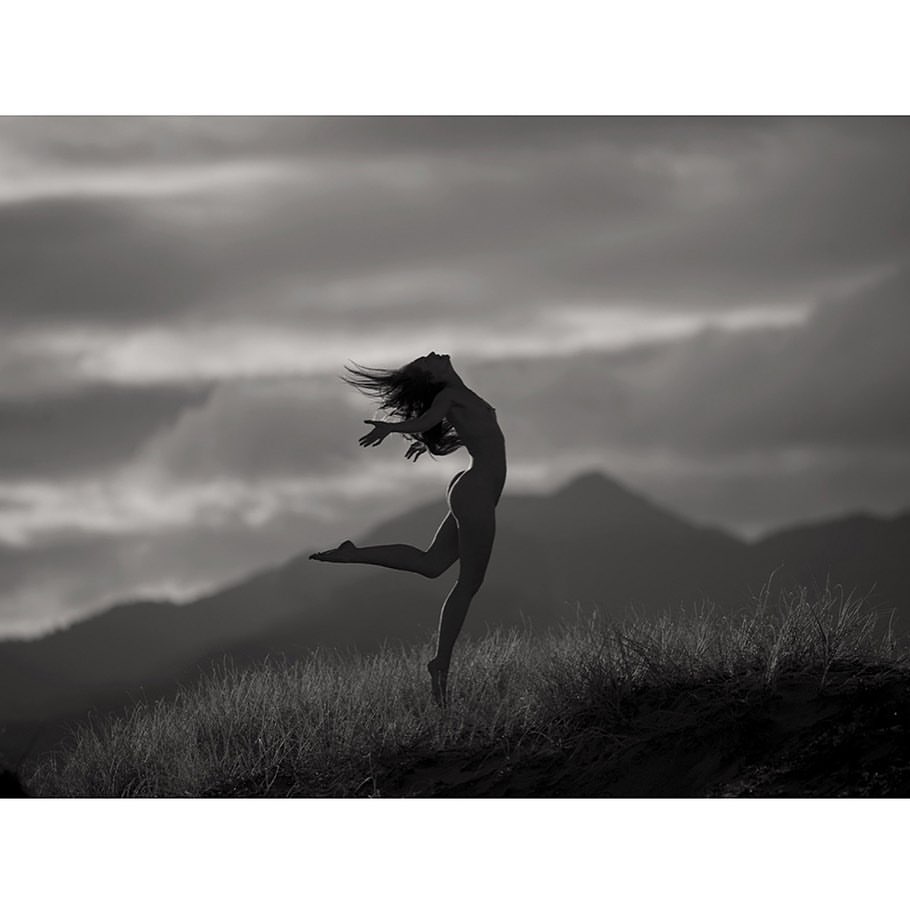 Silhouette of a woman leaping in a grassy landscape under a cloudy sky in a black and white artistic photograph.