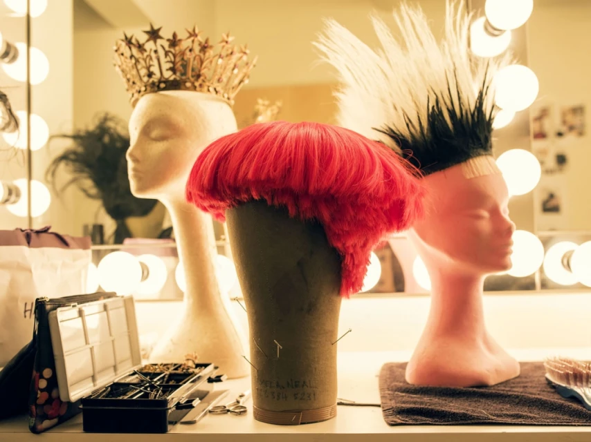 Wigs and headpieces sit on mannequin heads in a backstage dressing room, illuminated by warm vanity lights and professional tools.