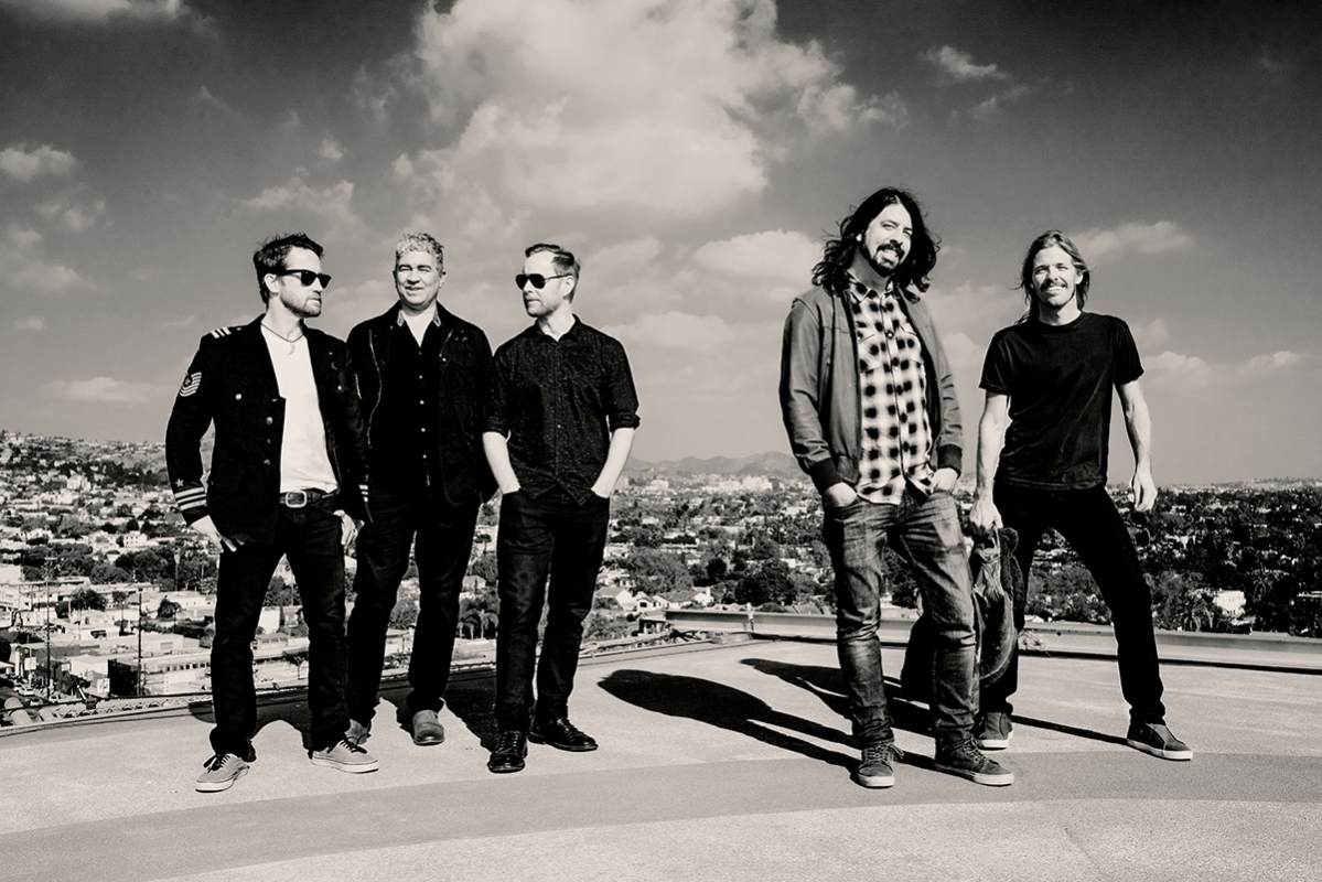 Black and white shot of five adult band members standing on a rooftop overlooking a cityscape with a bright cloudy sky above.