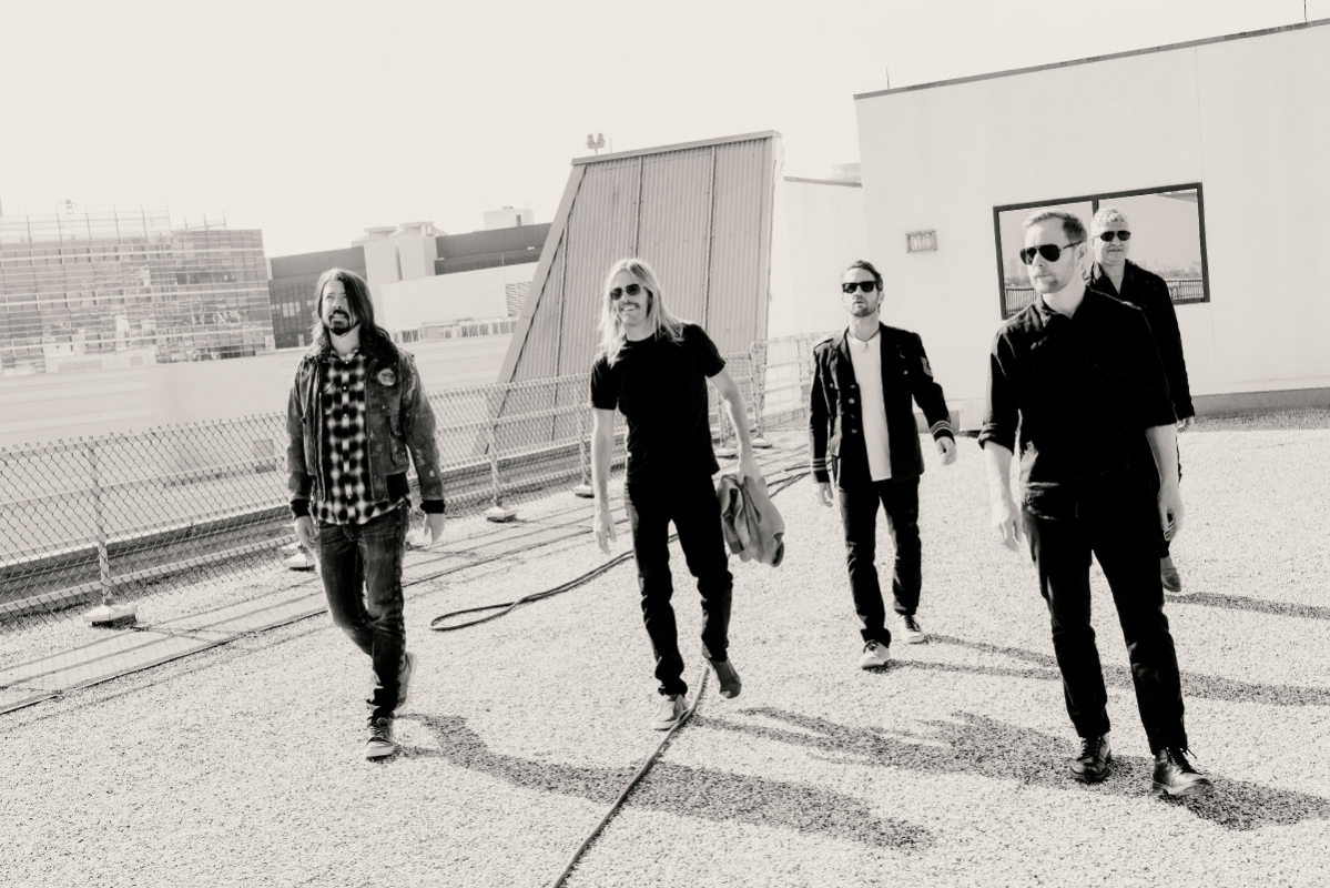A black and white shot of the band Foo Fighters, five adult males, walking outdoors on a rooftop against a bright sky.