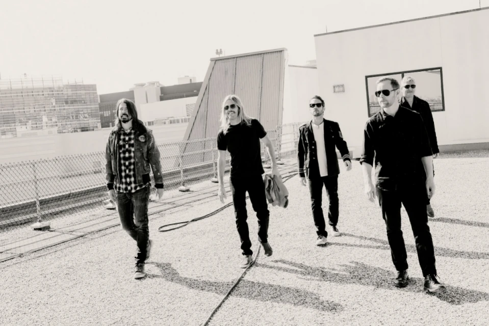 A black and white shot of the band Foo Fighters, five adult males, walking outdoors on a rooftop against a bright sky.