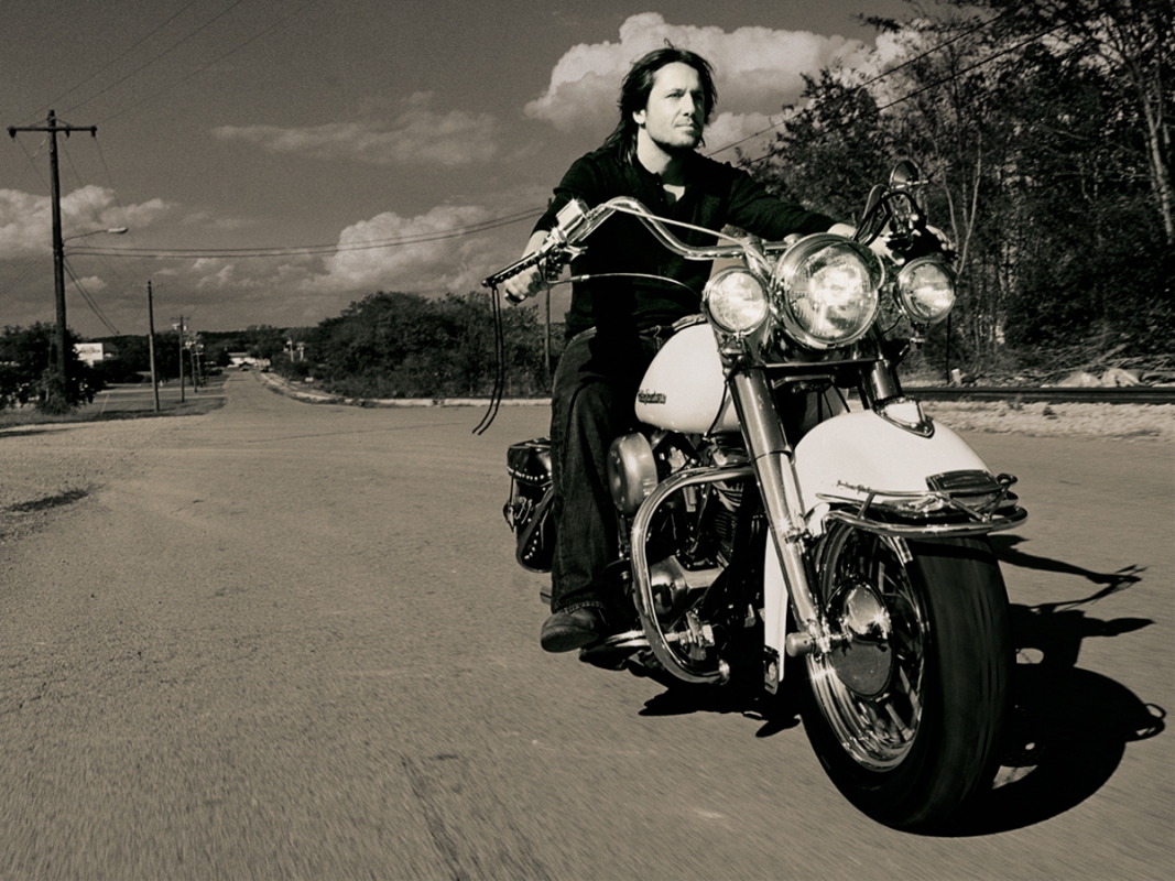 Monochrome shot of a man riding a vintage motorcycle on a rural road with power lines and trees, capturing a sense of freedom and movement.