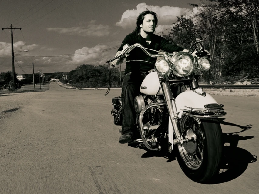 Monochrome shot of a man riding a vintage motorcycle on a rural road with power lines and trees, capturing a sense of freedom and movement.