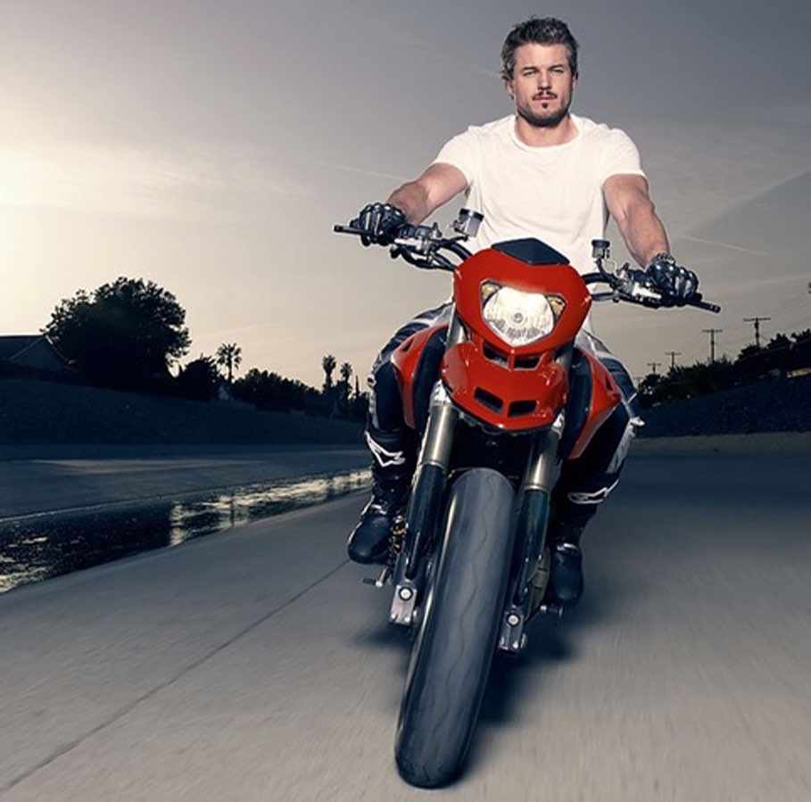 An adult male rides a red and black motorcycle on a paved road with a concrete barrier and hillside visible in the background.