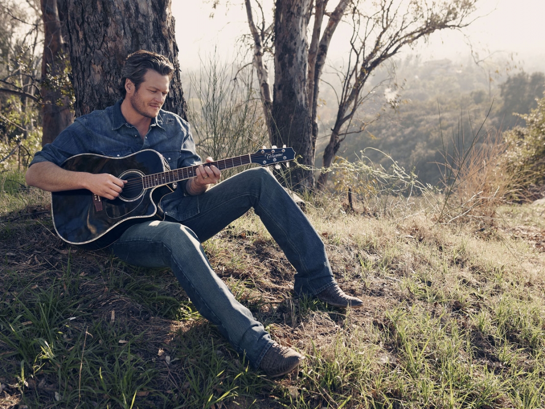 Adult man in blue denim shirt and jeans sits on grassy hillside playing black acoustic guitar near tree trunk on sunny day.