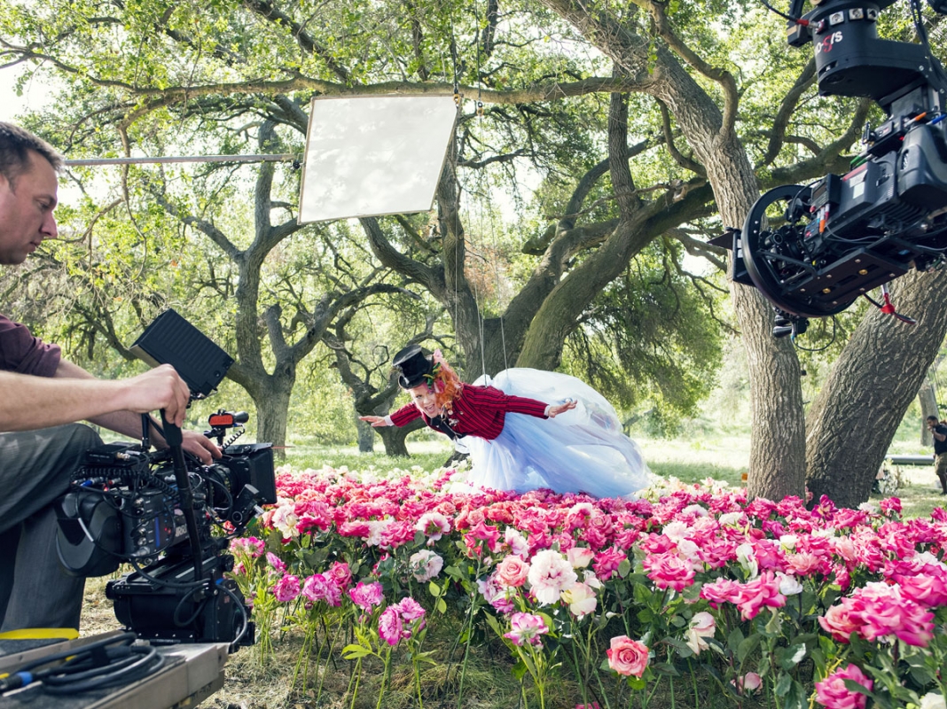 Behind-the-scenes shot of a child in costume in mid-air above a rose garden, with filming crew and equipment visible in the foreground.