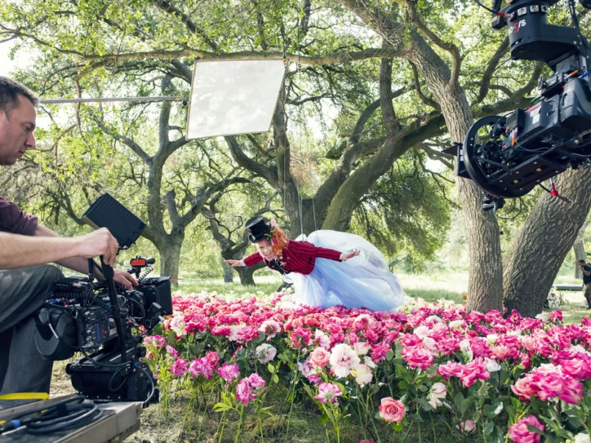 Behind-the-scenes shot of a child in costume in mid-air above a rose garden, with filming crew and equipment visible in the foreground.
