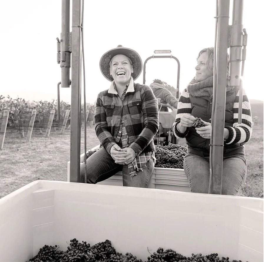 Monochrome photo of two women wearing casual clothing sitting on a vehicle surrounded by grapes at a vineyard during the daytime.