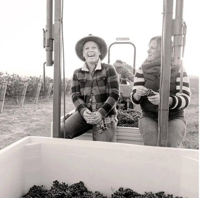 Monochrome photo of two women wearing casual clothing sitting on a vehicle surrounded by grapes at a vineyard during the daytime.