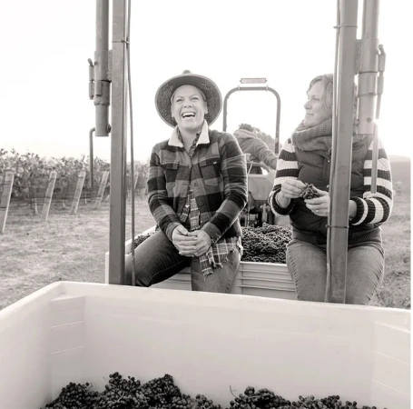 Monochrome photo of two women wearing casual clothing sitting on a vehicle surrounded by grapes at a vineyard during the daytime.