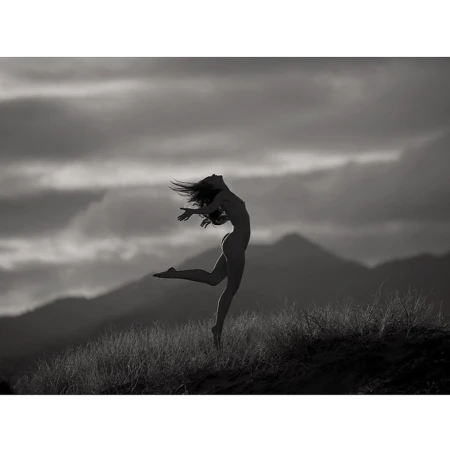 Silhouette of a woman leaping through the air over a grassy hill against a moody cloudy sky and mountain backdrop.