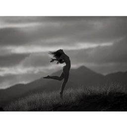Silhouette of a woman leaping through the air over a grassy hill against a moody cloudy sky and mountain backdrop.