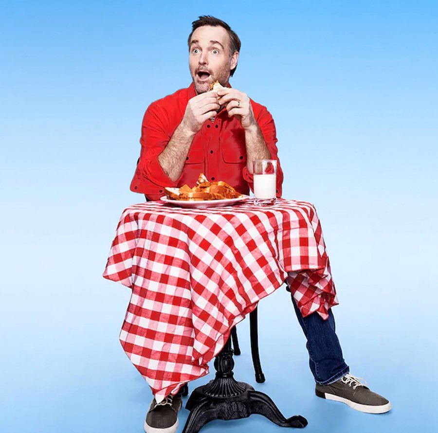 Adult male with an open mouth eating a sandwich at a table covered with a red and white checkered cloth in a blue photo studio.