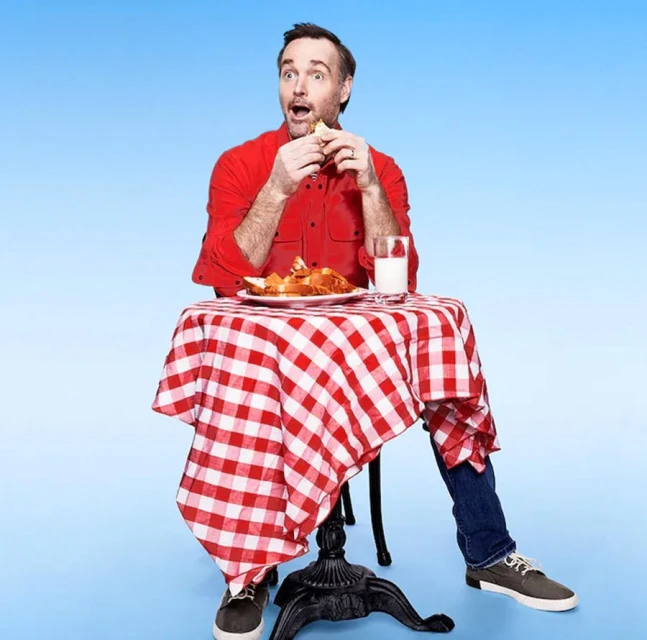 Adult male with an open mouth eating a sandwich at a table covered with a red and white checkered cloth in a blue photo studio.