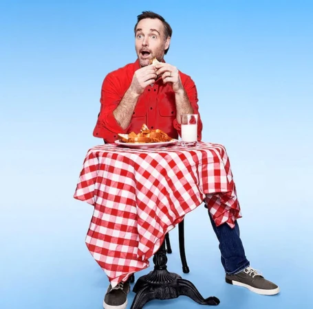 Adult male with an open mouth eating a sandwich at a table covered with a red and white checkered cloth in a blue photo studio.