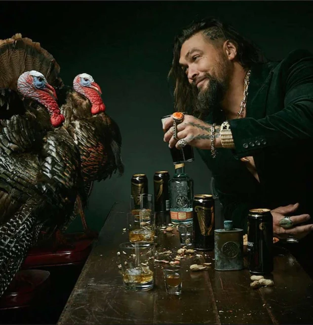 Jason Momoa poses with turkeys alongside cans and glasses of beer and a bottle of liquor on a wooden bar against a dark background.
