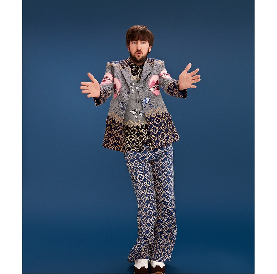 Full-body shot of a man in an ornate gray blazer and patterned pants against a blue background, striking a pose with open arms.