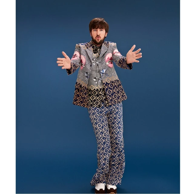 Full-body shot of a man in an ornate gray blazer and patterned pants against a blue background, striking a pose with open arms.