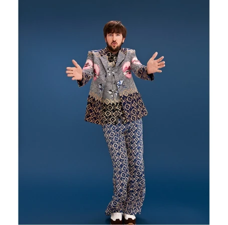 Full-body shot of a man in an ornate gray blazer and patterned pants against a blue background, striking a pose with open arms.