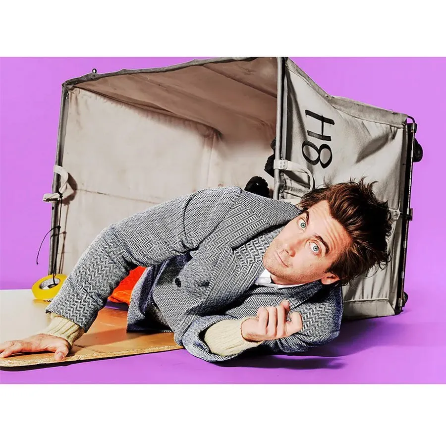 Adult male model lying beside a box prop on a purple backdrop in a studio setting, wearing a grey jacket and neutral sweater.