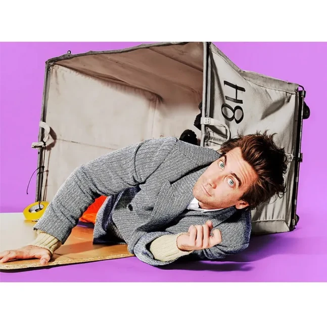 Adult male model lying beside a box prop on a purple backdrop in a studio setting, wearing a grey jacket and neutral sweater.