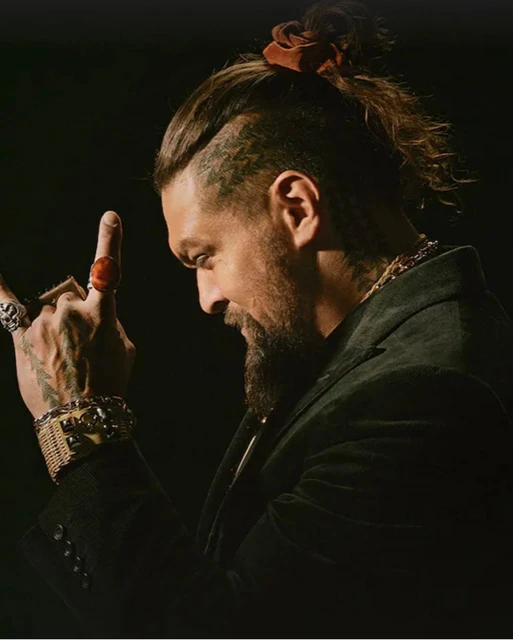 Side view of a stylish adult male with a beard, hair in a high bun, wearing rings, and a black jacket against a dark background.