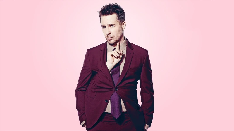 Portrait of a man in a maroon suit against a pink background, holding a finger to his lips, indicating silence or secrecy.