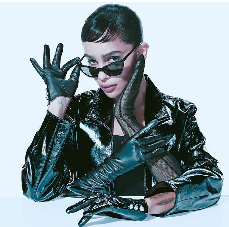 Stylish woman in a black leather jacket and gloves poses indoors with sunglasses, a sleek hairstyle, and a confident expression.