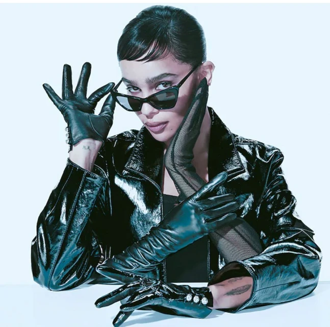 Stylish woman in a black leather jacket and gloves poses indoors with sunglasses, a sleek hairstyle, and a confident expression.